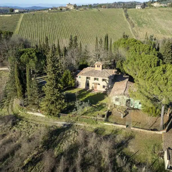 An aerial view reveals the Elegant Estate in the Hills of Greve in Chianti, featuring a rustic stone house surrounded by lush trees and greenery, with rolling hills, vineyards, and fields extending under a clear sky.